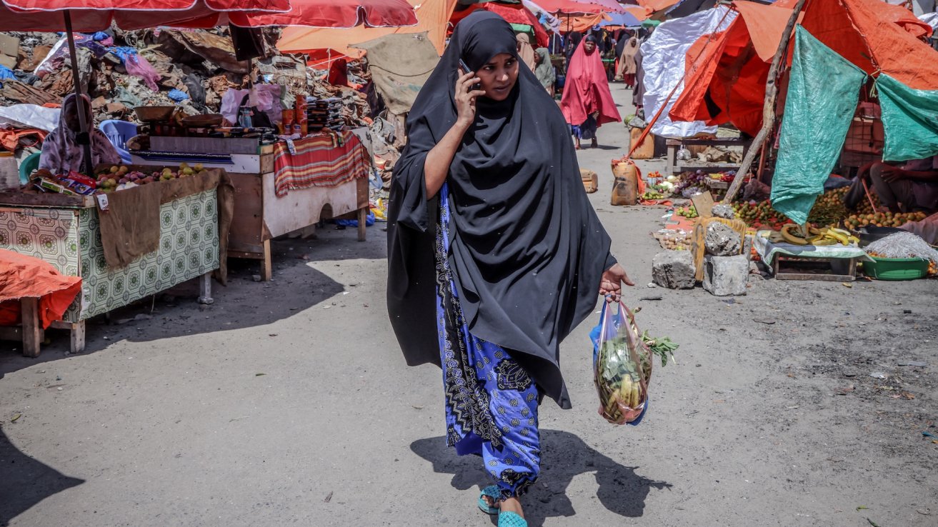 Woman in a black hijab walking through a bustling outdoor market while talking on a mobile phone, carrying a bag of vegetables.