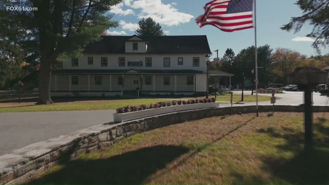 Front view of a white two-story building with a wraparound porch, black roof, and an American flag on a tall pole in the foreground; trees and flower bed surround the scene on a sunny day.