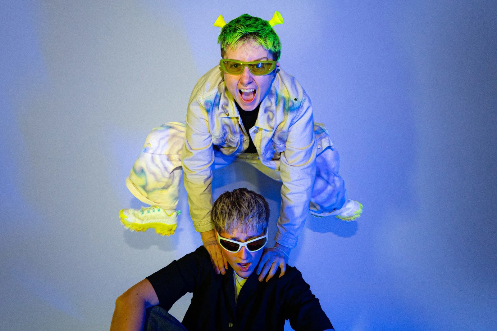 Two people in a blue-lit studio: one on the other’s shoulders wearing neon-green hair, yellow sunglasses, and a light jumpsuit, looking excited.