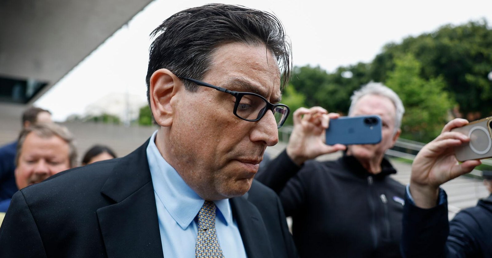 Man in a dark suit and glasses looks down as fans take photos with smartphones in the background.