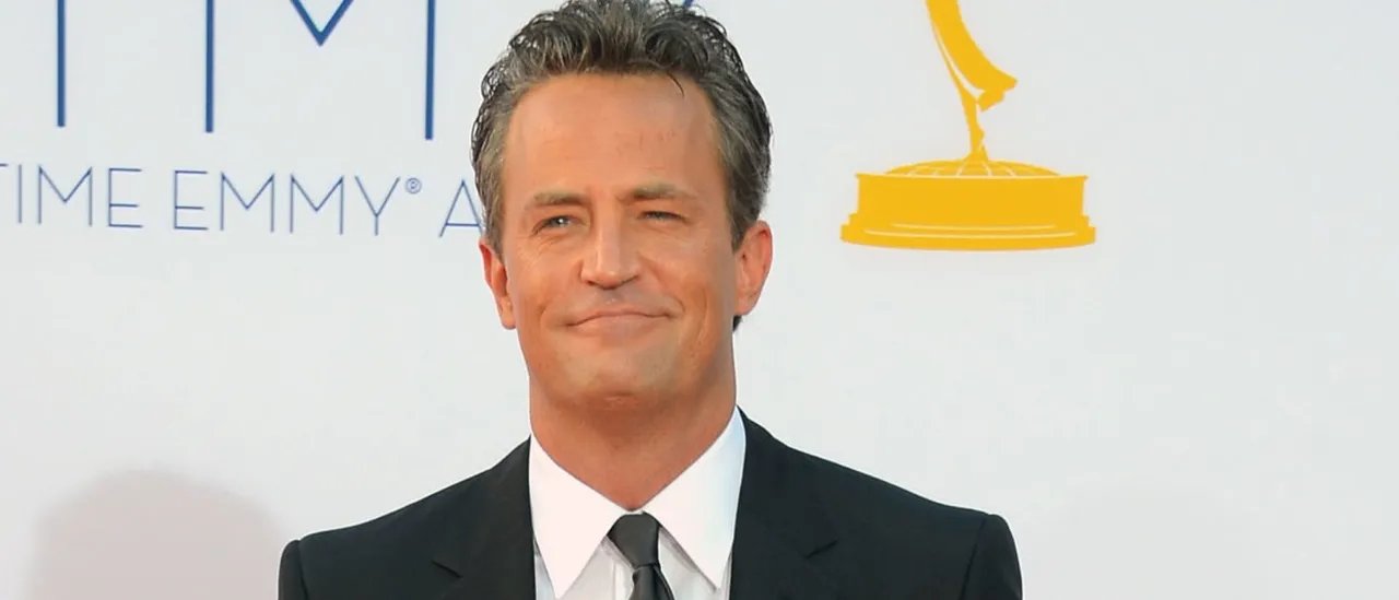Smiling man in a black suit and tie posing on a red carpet with an Emmy backdrop behind him.