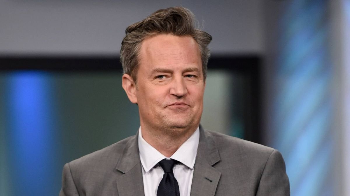 Portrait of a middle-aged man in a gray suit and black tie in a TV studio, smiling slightly.