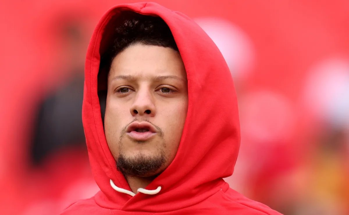 Close-up of a young man in a red hoodie speaking, neutral background blurred behind him
