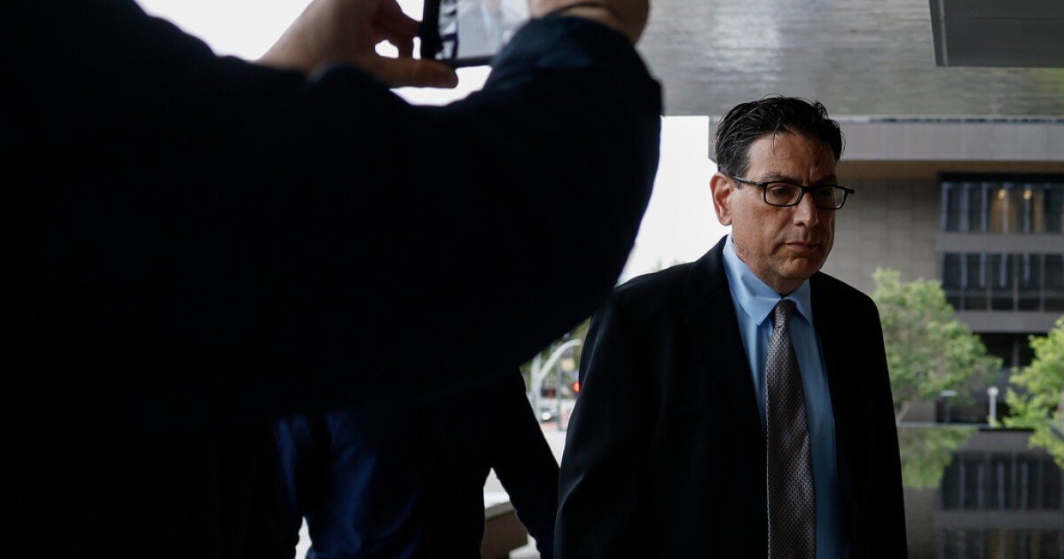 Businessman in a dark suit and glasses walks outside a modern office building, with a blurred person in the foreground holding a device nearby.