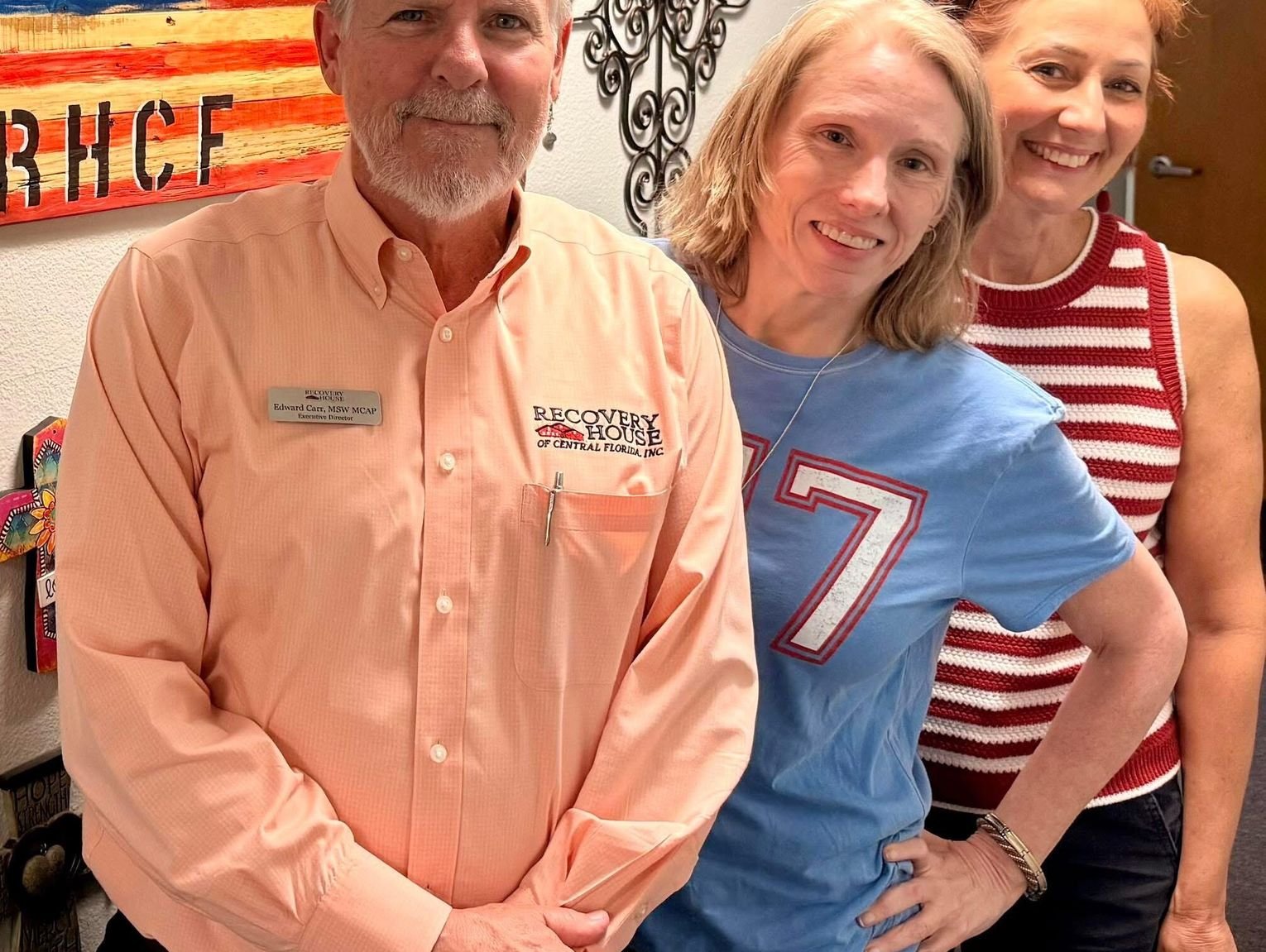 Three smiling adults posing indoors: man in peach shirt with name badge, woman in blue shirt with a large '7', and woman in a red-and-white striped sleeveless top.