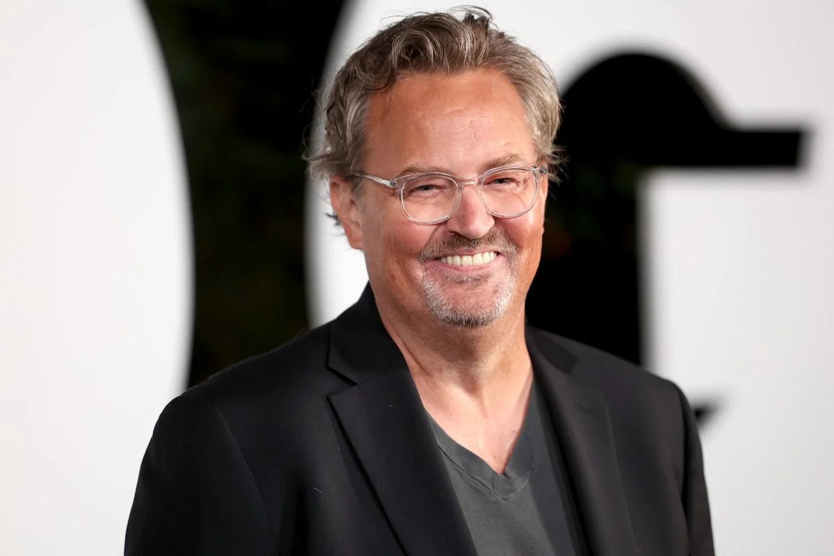 Smiling man with gray hair and glasses in a black blazer at a red-carpet event.