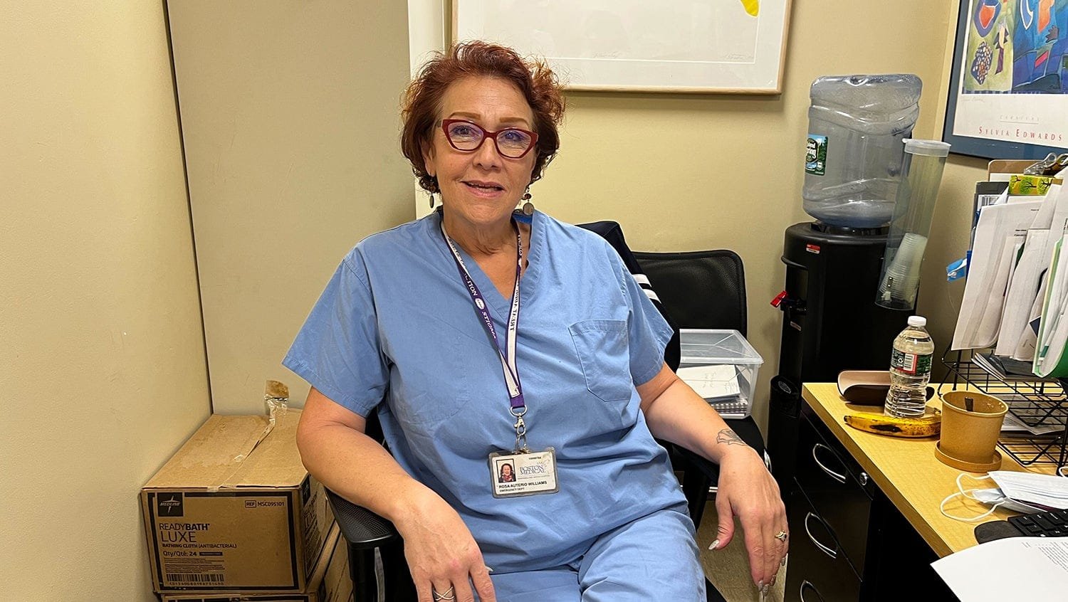 wp-header-logo-907png • Punkin's Patch Woman in blue scrubs with red glasses sits in an office chair, wearing an ID badge in a medical setting with files nearby.