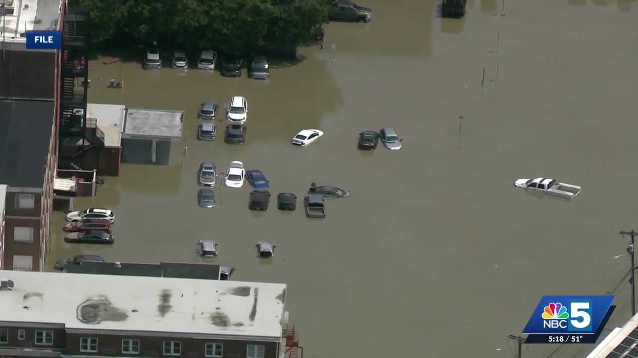 wp-header-logo-916png • Punkin's Patch Aerial view of a city street flooded, cars partially submerged in brown water with buildings on the left and a news channel watermark in the corner. Highlights widespread flooding in an urban area.