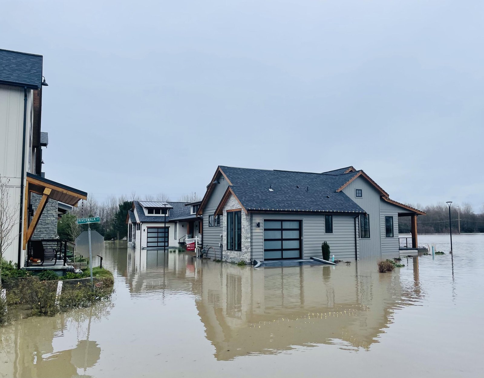 wp-header-logo-920png • Punkin's Patch Suburban street heavily flooded around modern houses, water up to door level, sign says 'Riverwalk' in the foreground, gray cloudy sky.