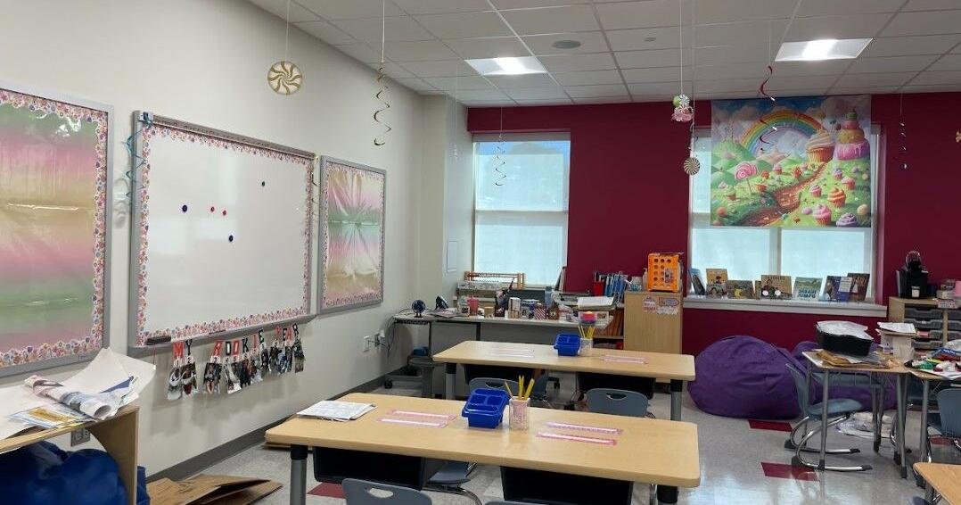 Bright classroom with whiteboards, desks, and colorful decorations; a purple beanbag and rainbow poster at the back wall.