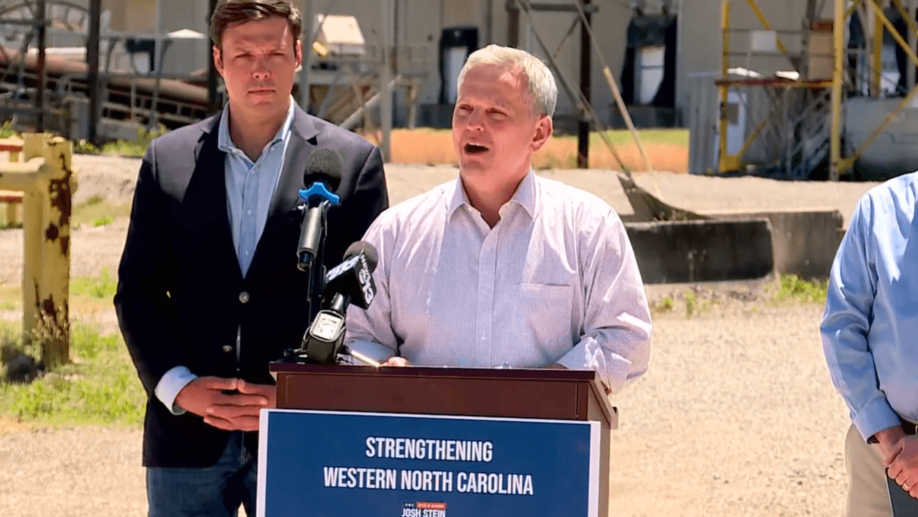 Two men at an outdoor construction-site press conference; one speaks at a podium with multiple microphones and a sign reading 'Strengthening Western North Carolina'.