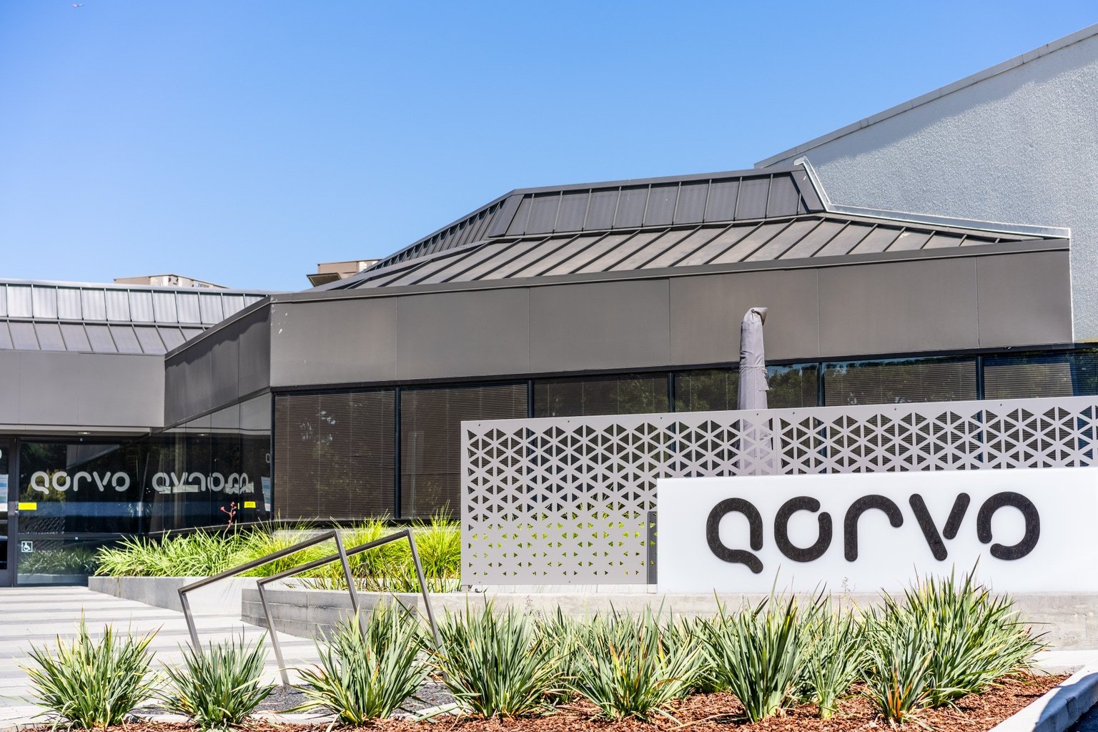 Modern office building exterior with a large Qorvo sign, metal panels, and decorative perforated screen, surrounded by landscaped plants.