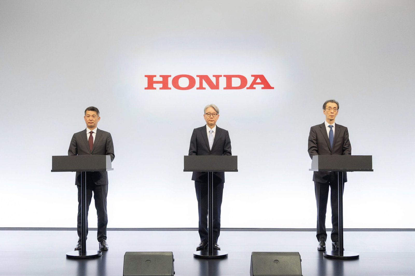Three men in dark suits stand at separate podiums on stage beneath a large red HONDA logo, presenting at a conference.