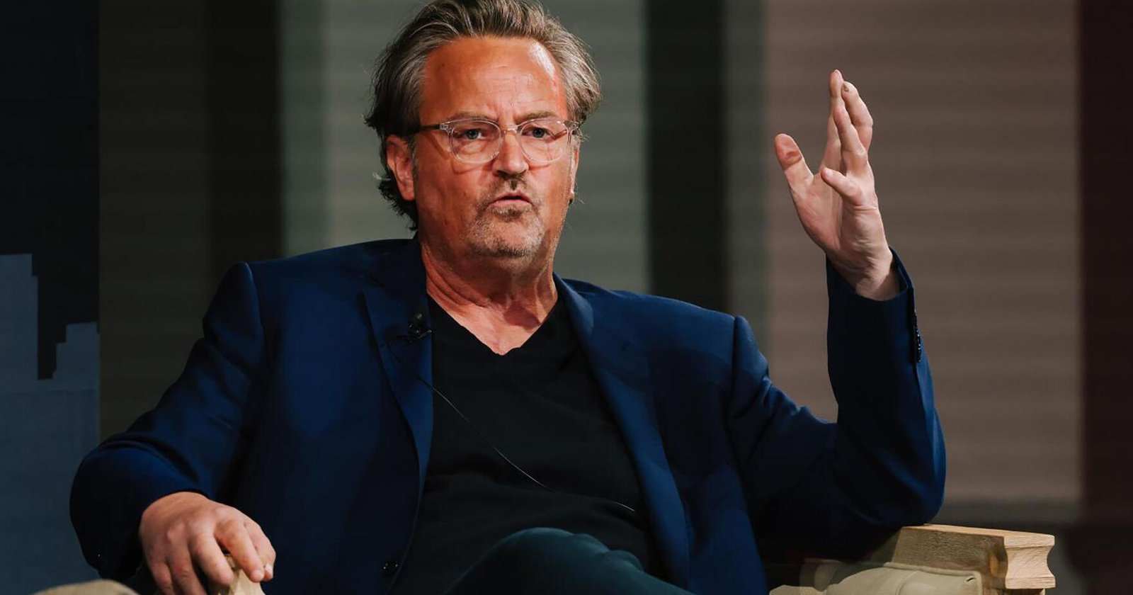 Man in a blue blazer and black shirt sits on a stage, gesturing with his right hand while speaking.
