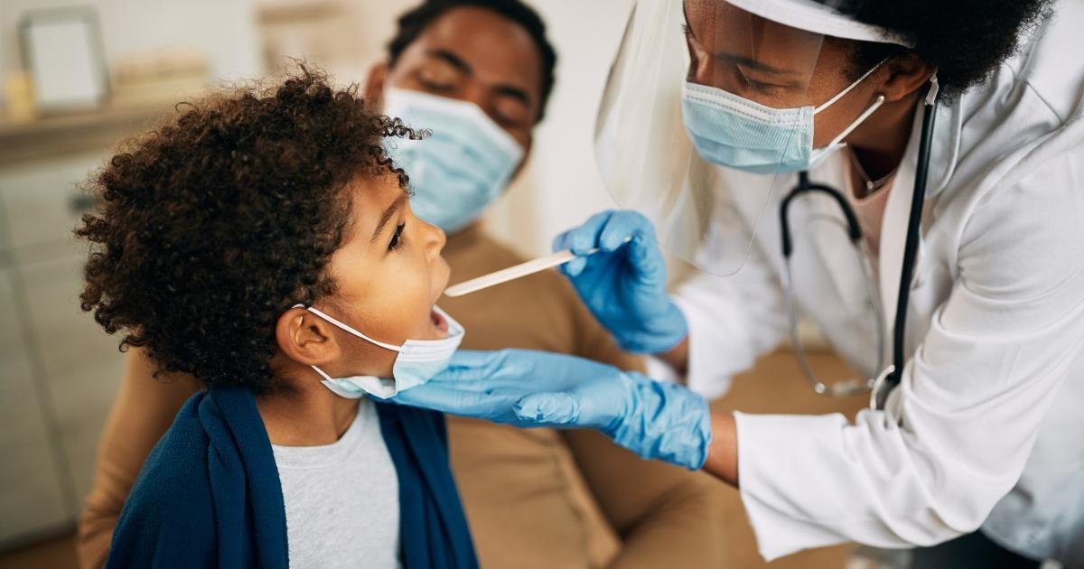 Healthcare worker in a mask and gloves swabs a child's mouth during a medical test, with a parent in the background approvingly watching.