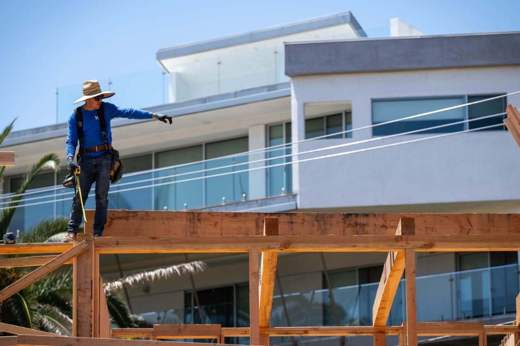 Construction worker wearing a straw hat and blue shirt standing on a wooden framework of a building under construction, pointing ahead.
