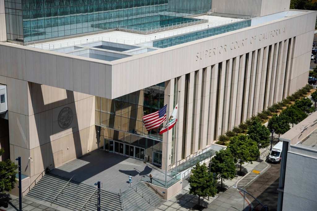 Aerial view of a modern courthouse with tall beige columns, glass entrance, and U.S. and California flags outside.