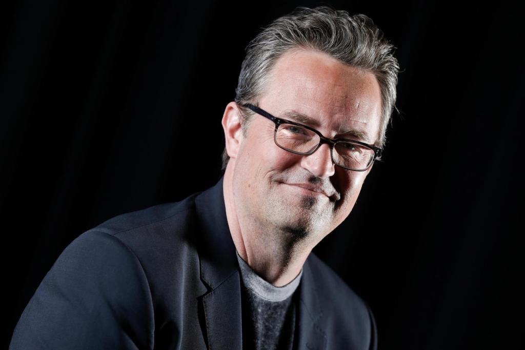 Smiling man with dark hair and glasses in a dark studio setting, wearing a navy blazer.