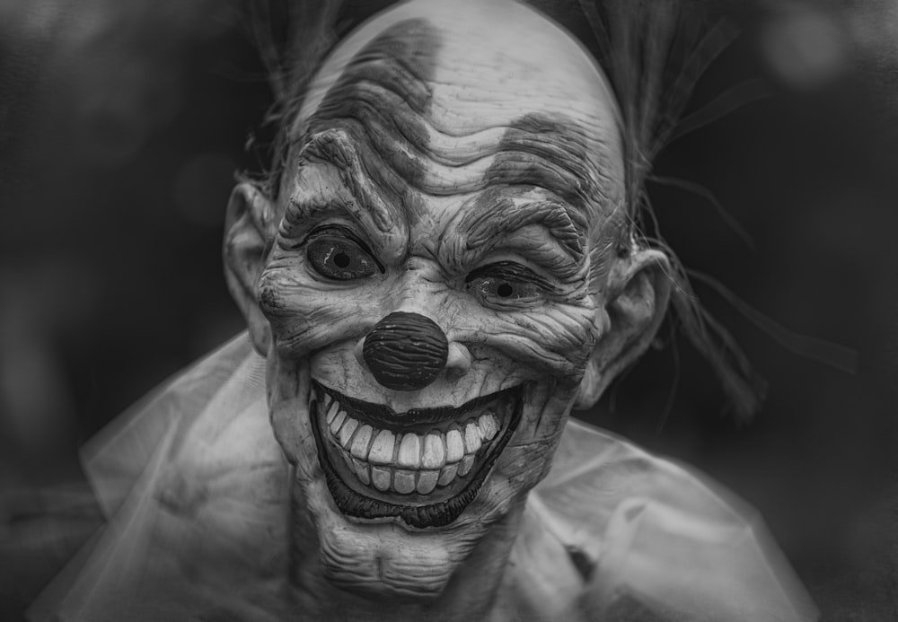 Creepy clown with a wide, toothy grin and exaggerated facial wrinkles, pale makeup, and a rounded nose in a black-and-white portrait.