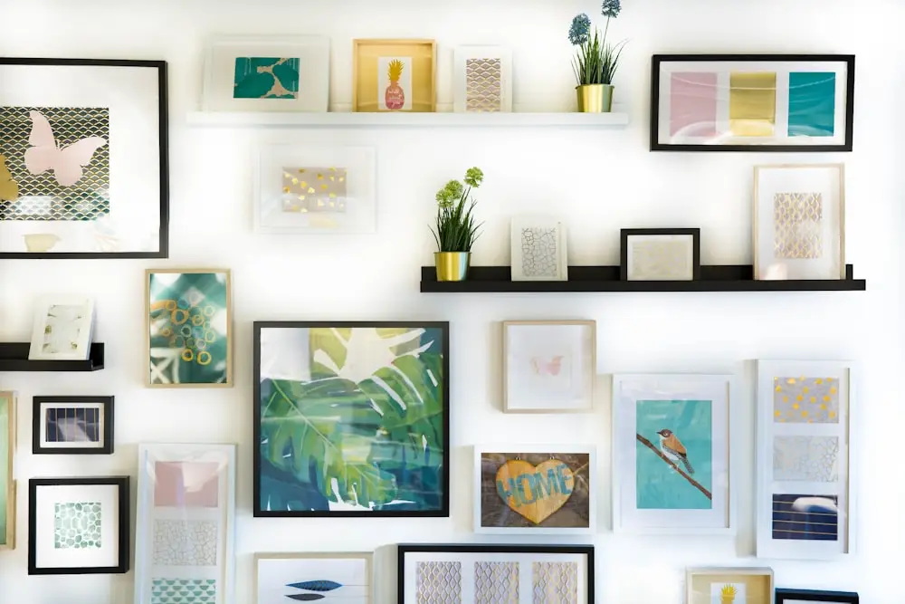 Wall gallery of assorted framed art and small plants on white and black shelves, colorful abstract pieces and photos.